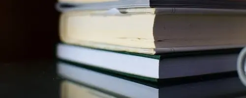 Close-up of a stack of three books placed on a reflective surface, with the edges of the pages and covers visible. The books are partially open, showing slightly bent pages.