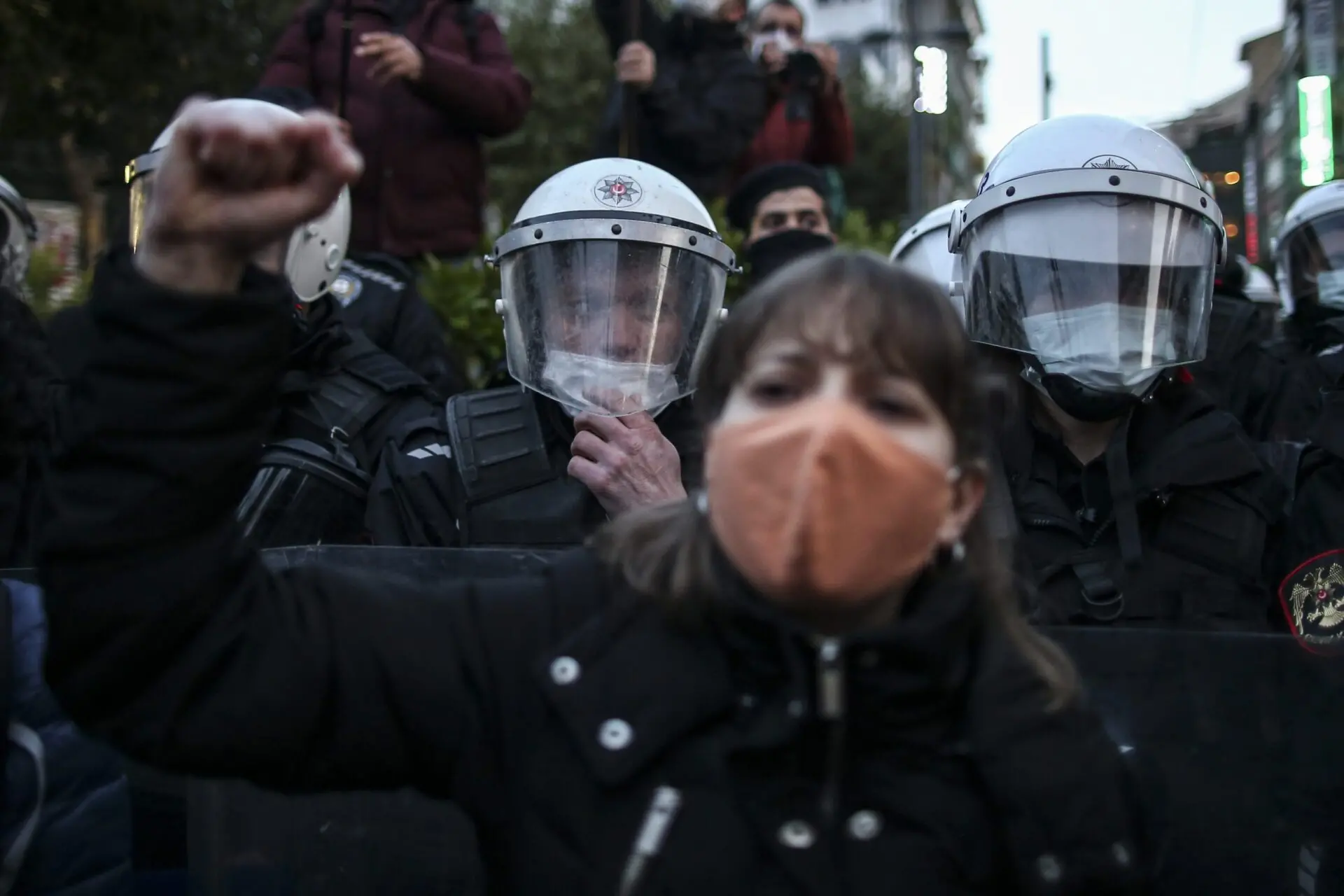 A woman wearing a face mask raises her fist in protest in front of a line of police officers in riot gear and helmets during a demonstration.