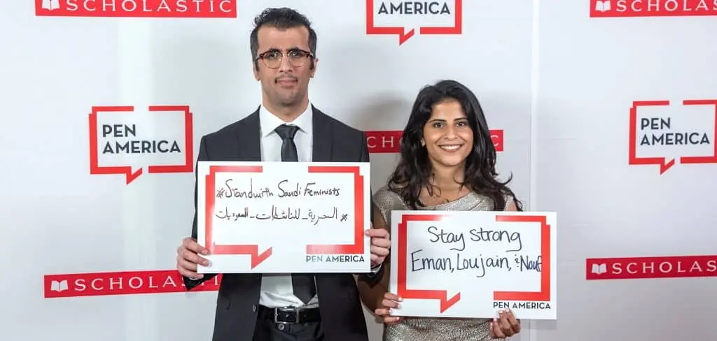 Two people stand in front of a backdrop with PEN America and Scholastic logos, holding signs supporting Saudi feminists and expressing solidarity. The man wears a suit and glasses; the woman has long dark hair and wears a dress.