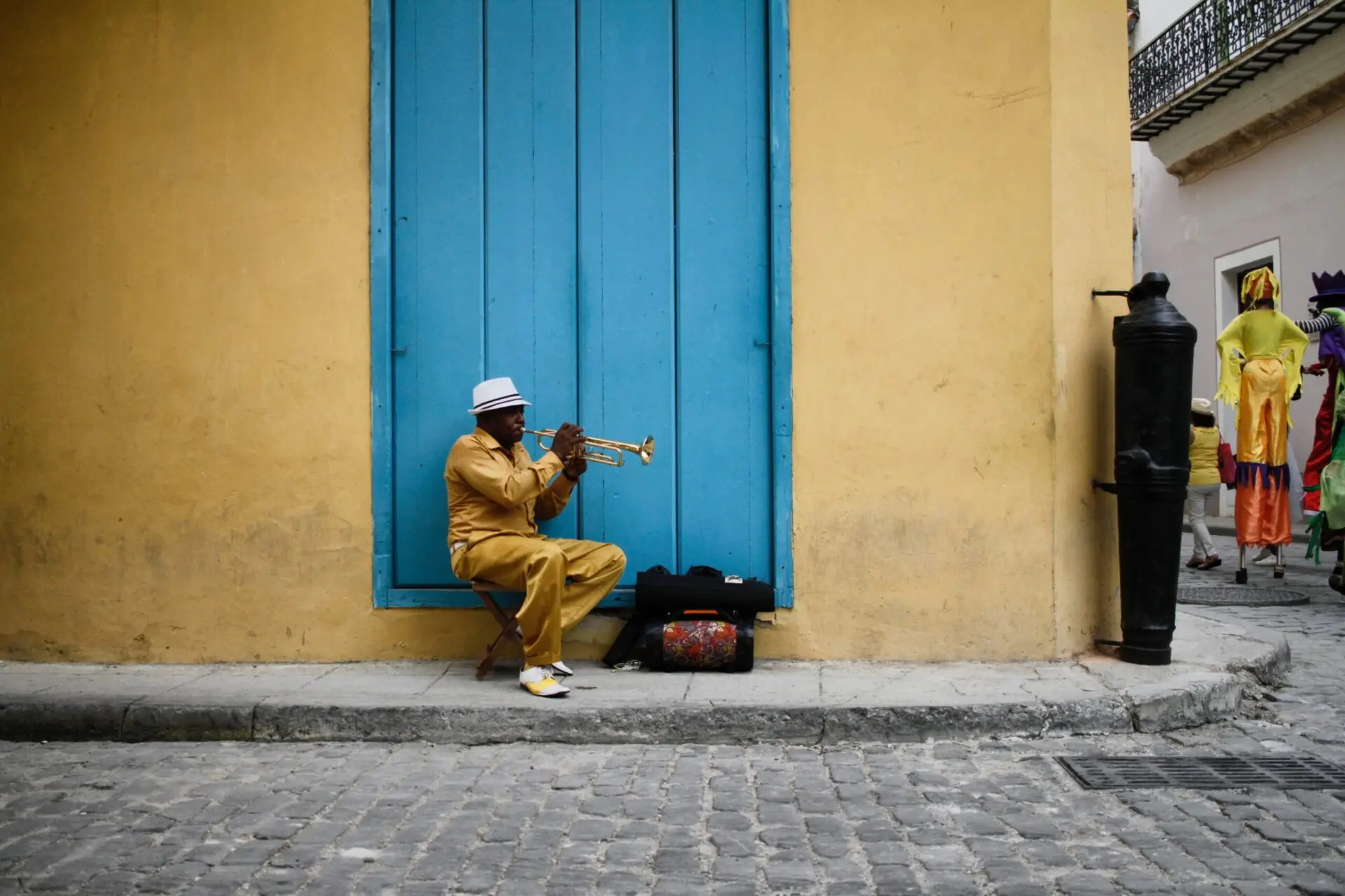 A man in yellow clothes and a white hat sits on a stool, playing a trumpet in front of a large blue door set in a yellow wall. His instrument case and a bag are on the ground next to him.