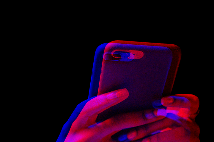A close-up of a hand holding a smartphone with a black background. The image has a double exposure effect with red and blue lighting, creating a blurred, futuristic appearance.