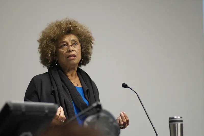 A woman with curly hair and glasses speaks at a podium. She wears a black jacket and scarf over a blue top, gesturing with her hands. A microphone and a metal tumbler are visible in front of her.
