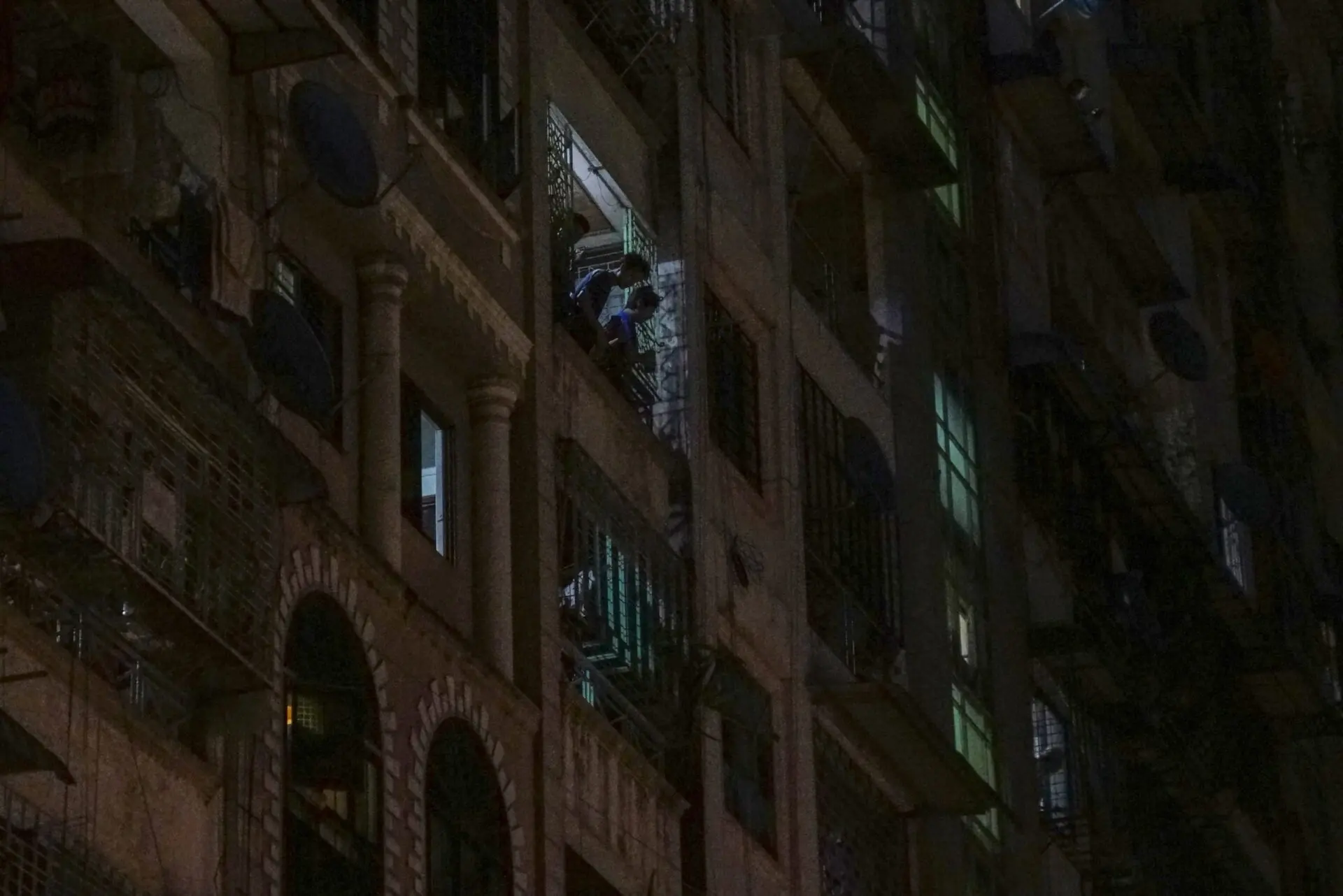 A dimly lit apartment building at night, with many windows and balconies, some illuminated. A person is visible on one brightly lit balcony, while other apartments are mostly dark.