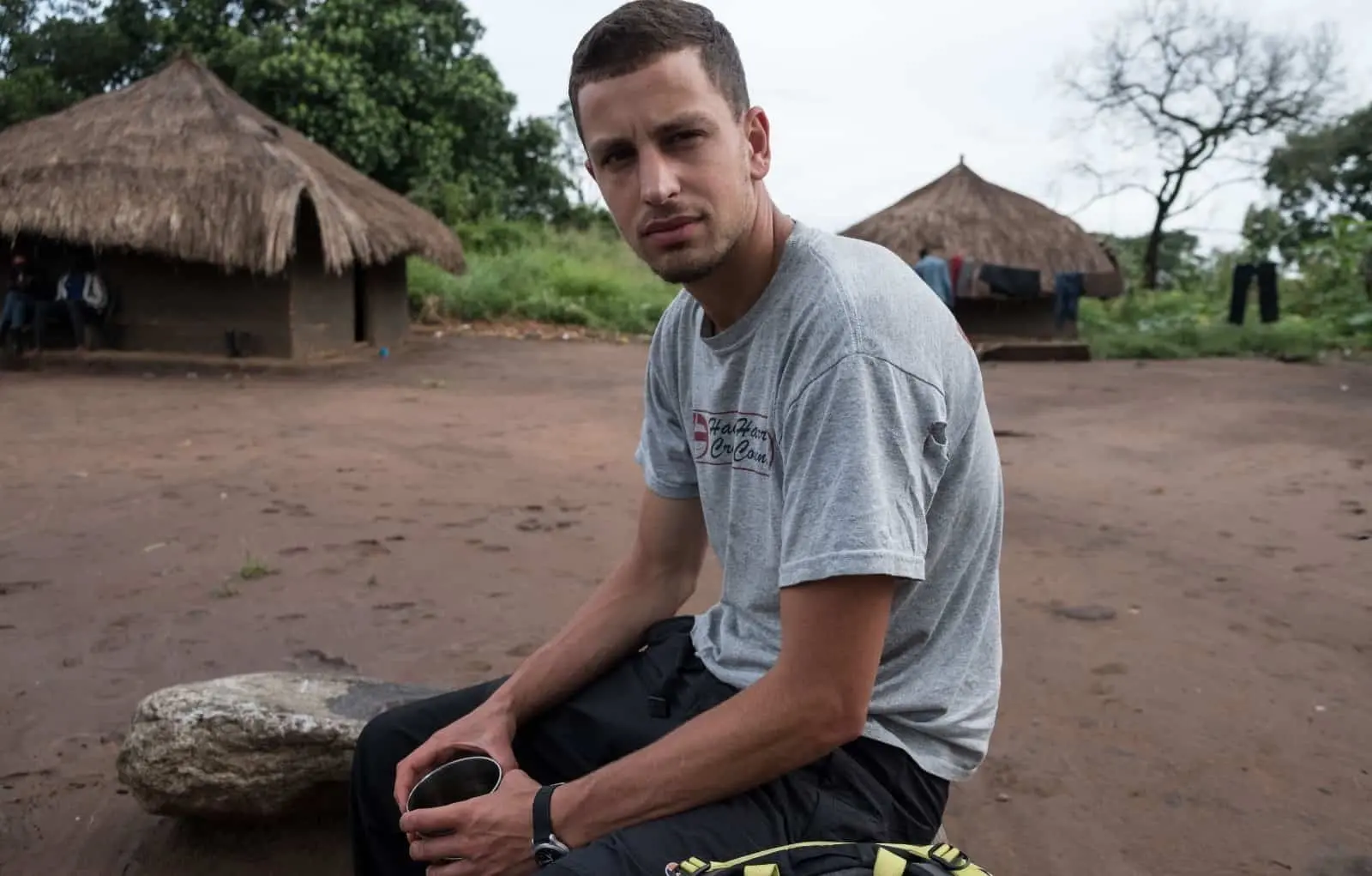 A man in a gray t-shirt sits on a rock holding a metal cup, with traditional thatched-roof huts and trees in the background, on a dirt ground in a rural setting.