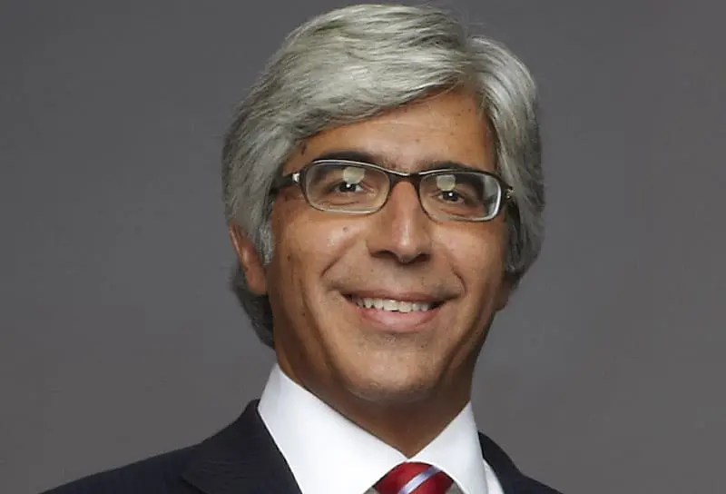 A middle-aged man with gray hair wearing glasses, a dark suit, a white shirt, and a red striped tie, smiling in front of a plain gray background.