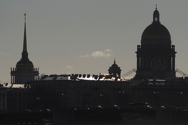Silhouette of city buildings with a tall spire and a large dome against a dusky sky, some rooftops reflecting light, and faint clouds in the background.