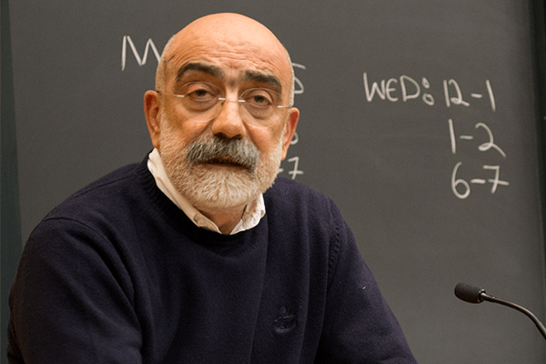 An older man with a bald head, gray beard, and glasses speaks at a podium in front of a chalkboard with handwritten notes and times listed for Wednesday.
