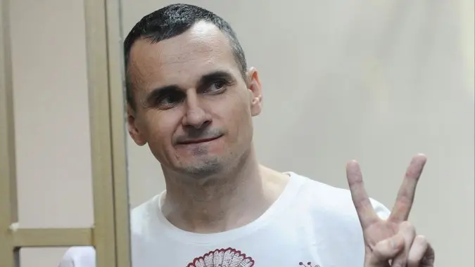 A man with short dark hair, wearing a white shirt, stands behind a glass panel and smiles while making a peace sign with his right hand.