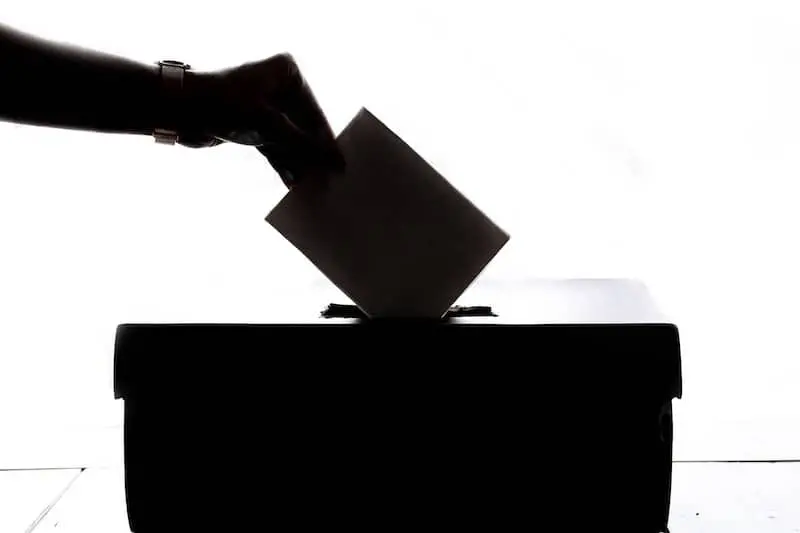 A silhouetted hand places a ballot into a box against a white background, symbolizing voting or an election process.