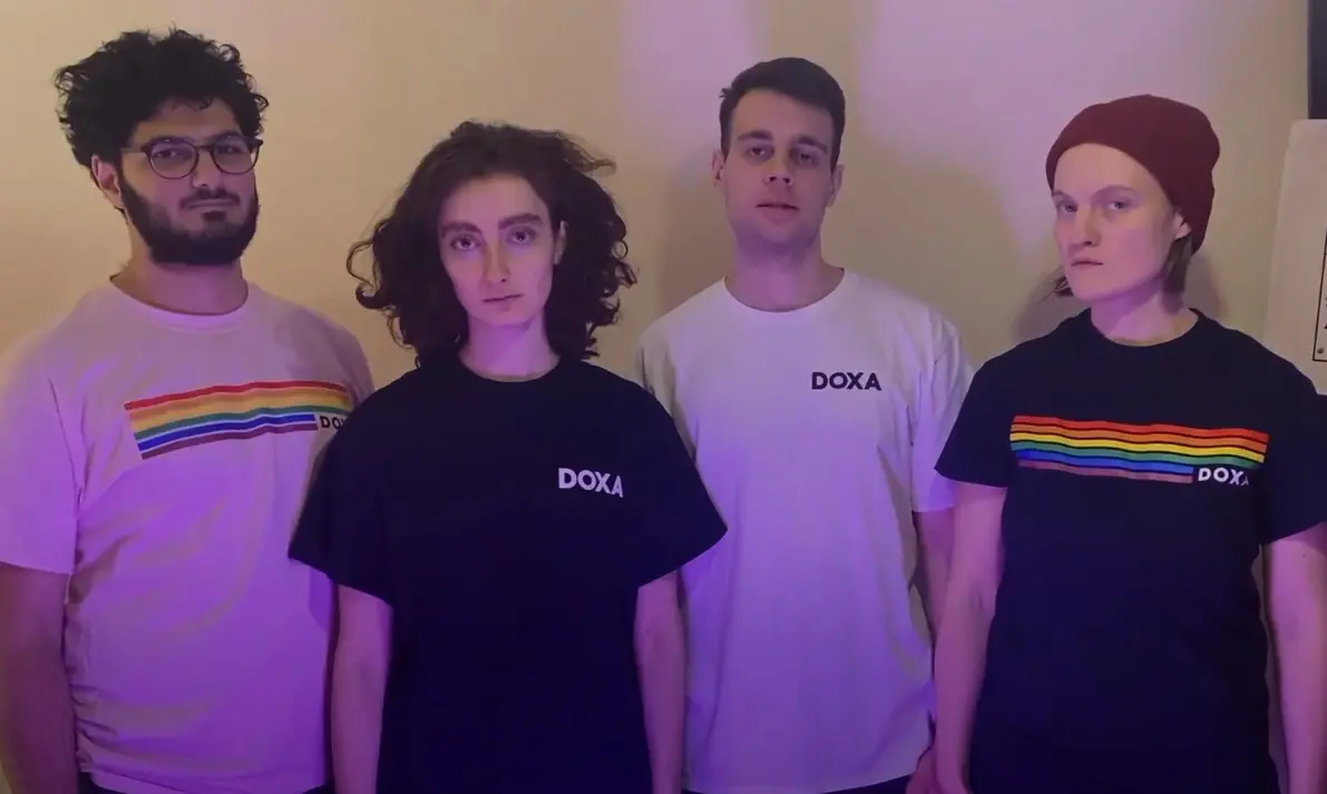Four people stand side by side against a beige background, wearing DOXA t-shirts; two shirts have rainbow stripes, and two are plain. The group looks directly at the camera with neutral expressions.