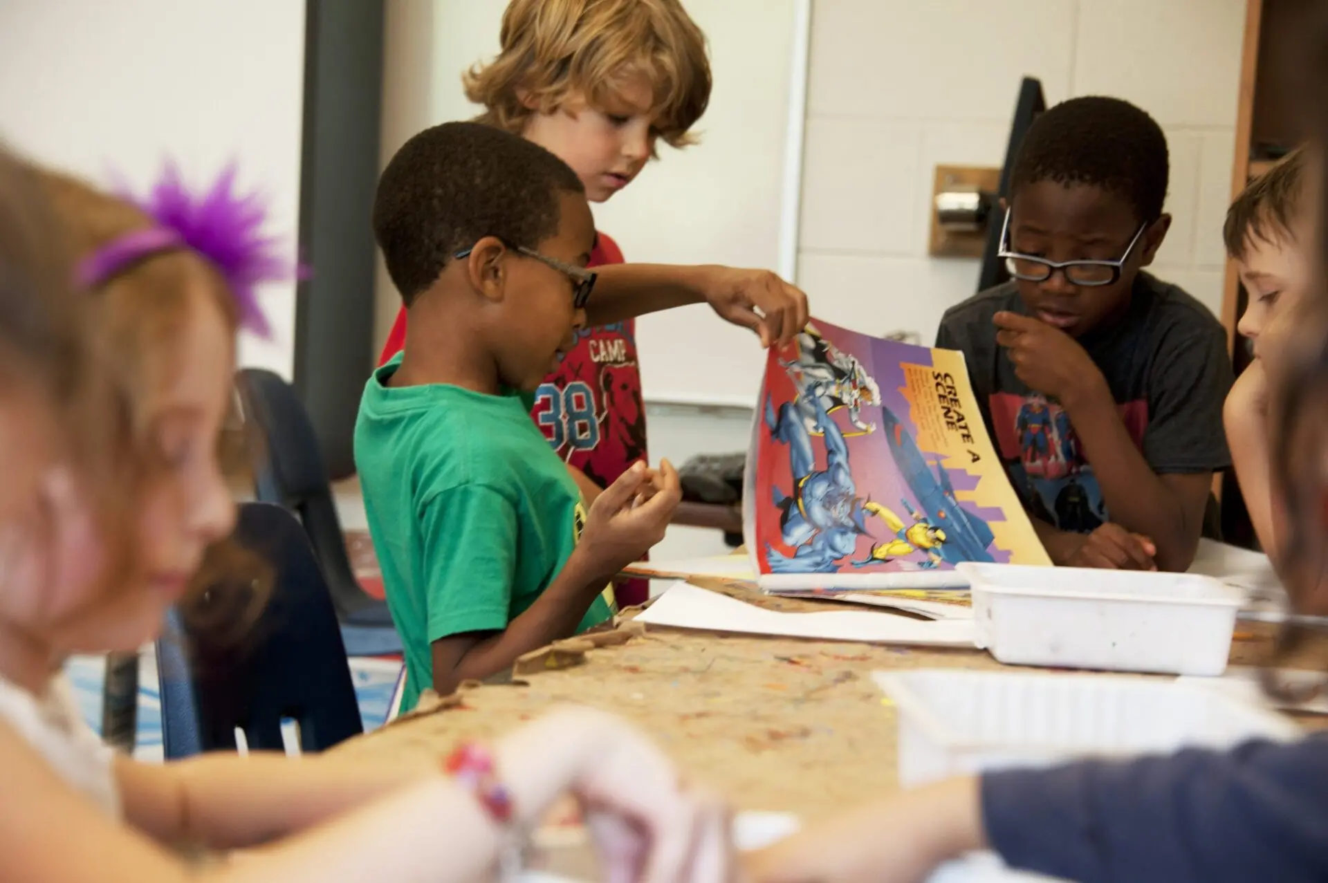 Several children sit and stand around a table in a classroom, working on arts and crafts. Two boys look at a colorful comic book together while others focus on their own projects.