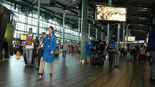 Busy airport terminal with travelers walking and pulling suitcases. Two staff members in blue uniforms walk in the foreground. Large windows and information screens are visible in the bright, spacious hall.