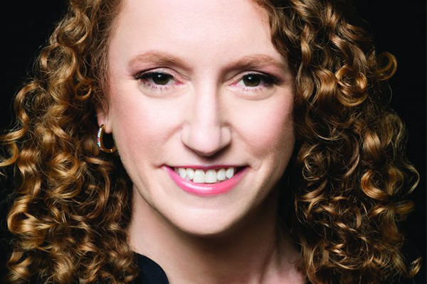 A woman with fair skin and curly, reddish-brown hair smiles at the camera. She is wearing light pink lipstick, subtle makeup, and a small hoop earring, posed against a dark background.