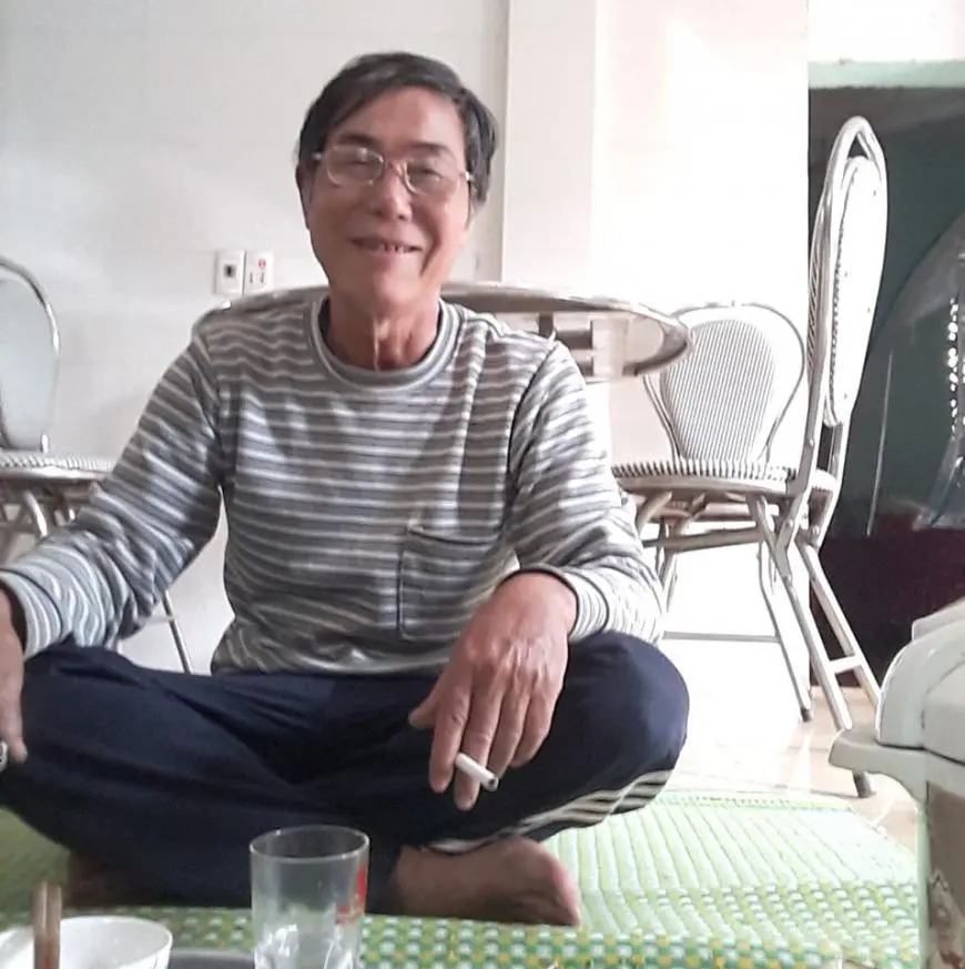 An older man wearing glasses and a striped shirt sits cross-legged on a mat indoors, smiling at the camera. There are empty glasses and bowls in front of him, with chairs and a table in the background.