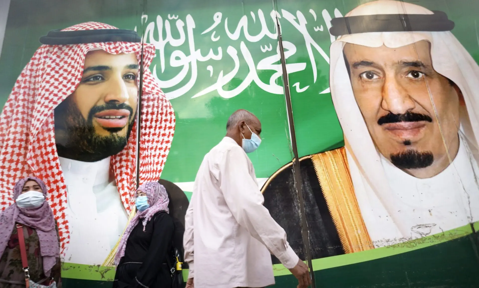 Three people wearing face masks walk past a large mural featuring two Saudi leaders in traditional attire, with the Saudi Arabian flag and Arabic script in the background.