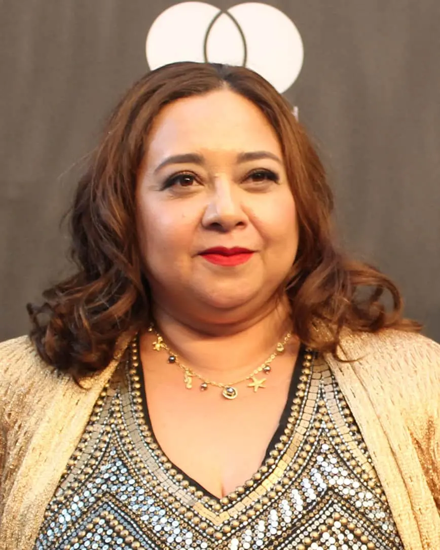 A woman with wavy brown hair wears a gold and black sequined top, a gold necklace with charms, and a gold jacket. She is standing in front of a dark backdrop with a white circular logo.