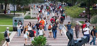 A large group of students walks along a wide, outdoor brick pathway lined with greenery on a college campus during the day. Some carry backpacks and talk with friends. The atmosphere is lively and busy.