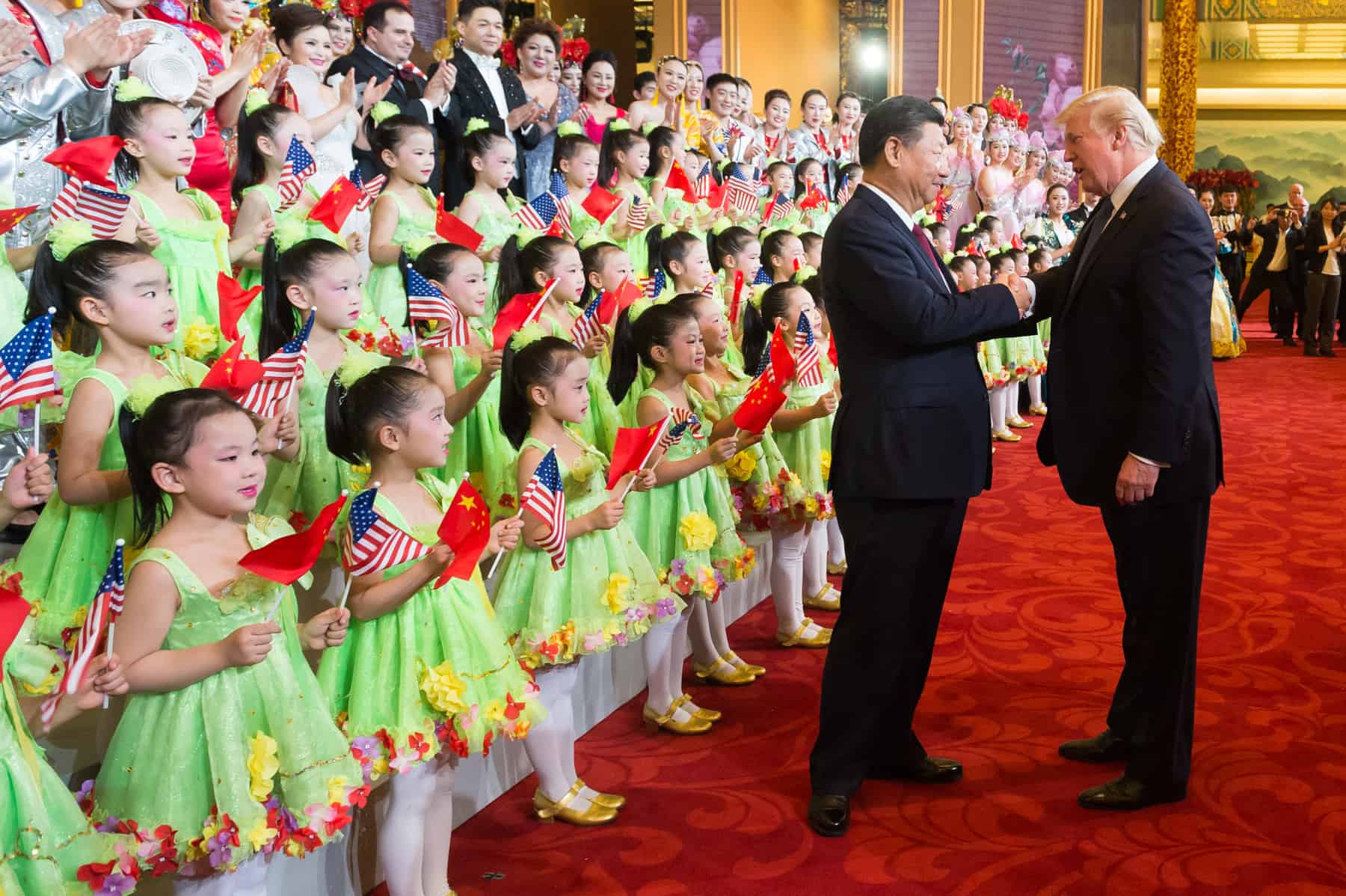 A group of young girls in green dresses holding Chinese and U.S. flags stand in rows, while two men in suits shake hands on a red carpet inside an ornate hall. Adults and performers are in the background, smiling.