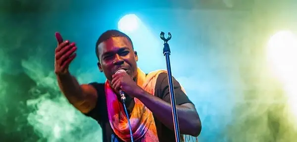 A man passionately sings or raps into a microphone on stage, with colorful lights and smoke in the background, wearing a scarf draped over one shoulder and gesturing with his hand.
