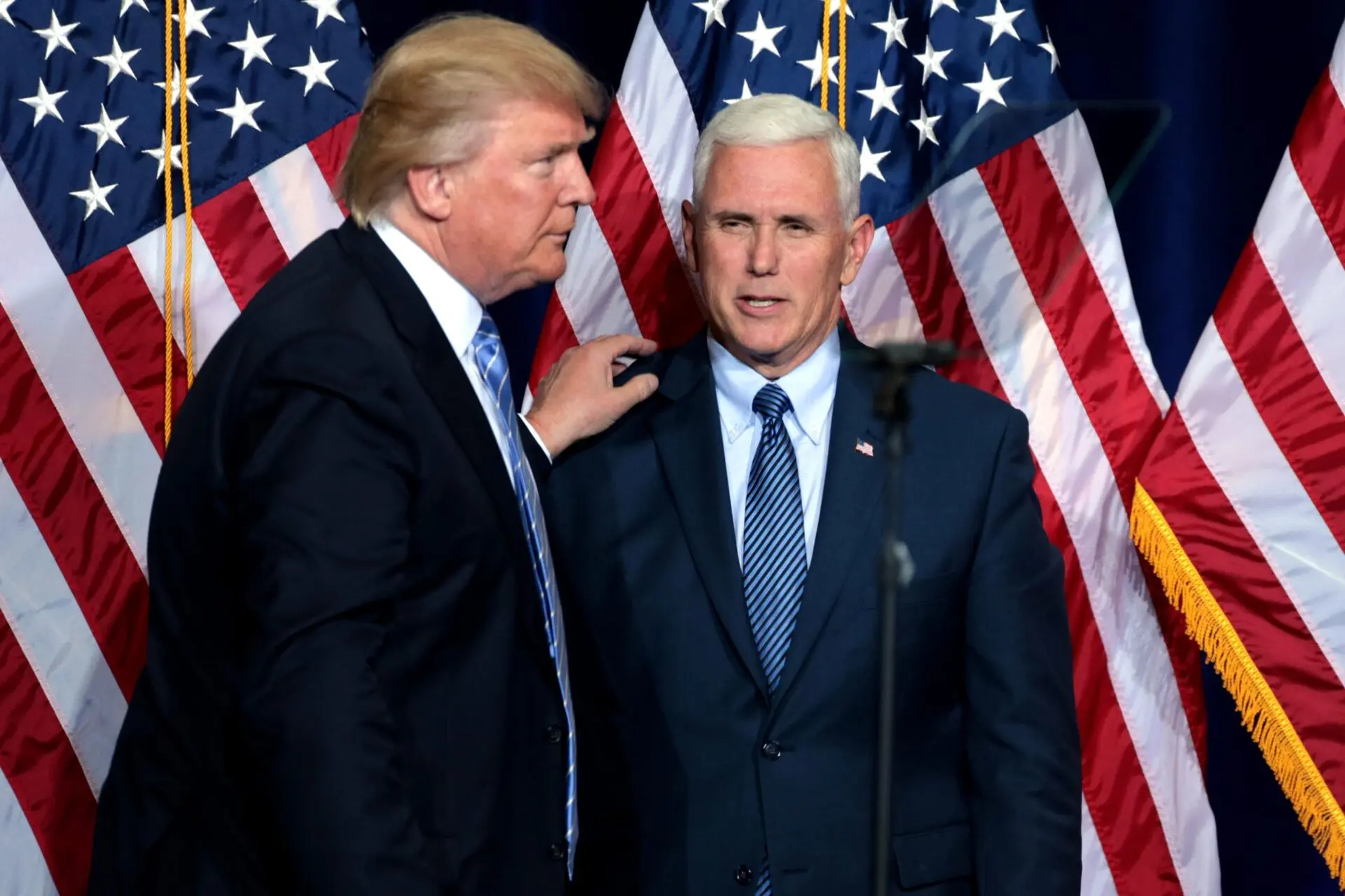 Two men in suits stand in front of several American flags; one man with blond hair is placing his hand on the other mans shoulder as they face each other.