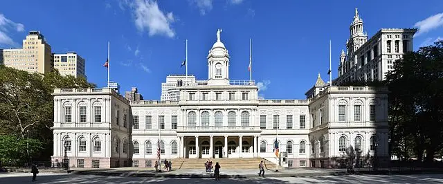 A wide view of New York City Hall, a historic white building with a central dome and clock, surrounded by trees and tall skyscrapers under a bright blue sky with scattered clouds.