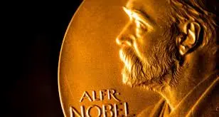 Close-up image of a Nobel Prize medal featuring a side profile of Alfred Nobel with his name partially visible engraved on the medal. The medal has a golden color.