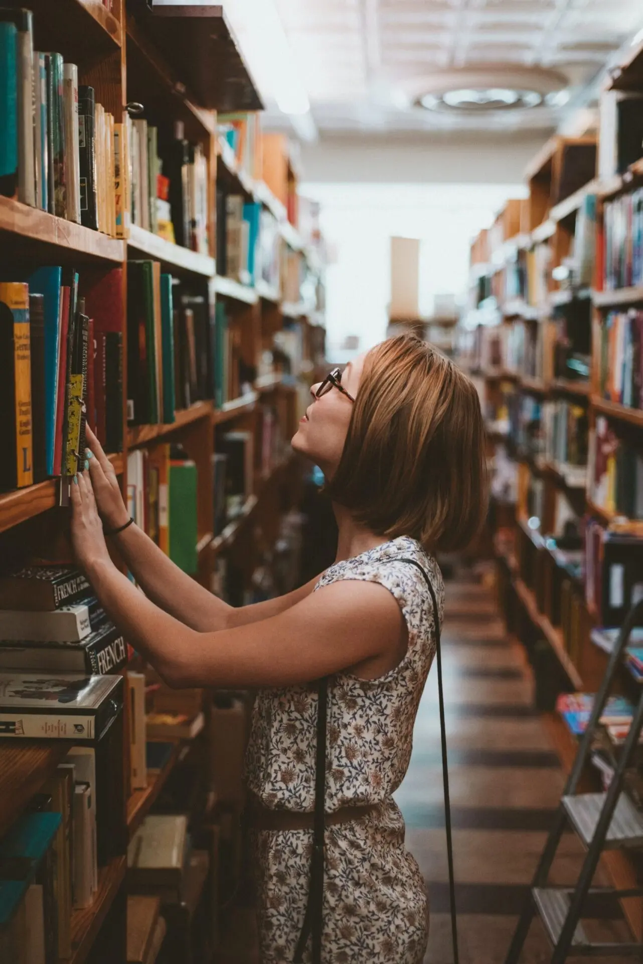 A woman wearing glasses and a floral dress browses books on a tall shelf in a library or bookstore, reaching up to select a book. Bookshelves filled with books line both sides of the narrow aisle.