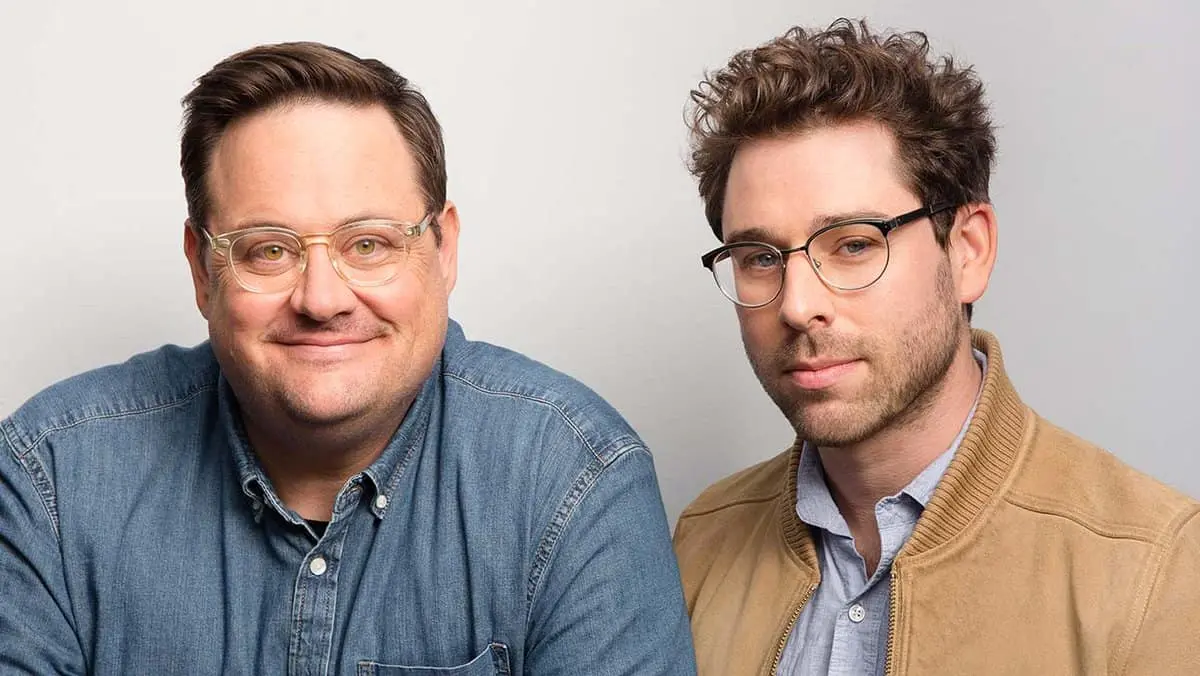 Two men with glasses pose together in front of a plain light background. The man on the left wears a denim shirt and smiles, while the man on the right has curly hair, a tan jacket, and a neutral expression.
