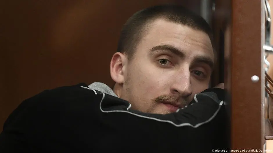 A man with short hair and a mustache leans his head and arms against a wooden surface, looking thoughtfully at the camera. He is wearing a dark-colored top with light trim.