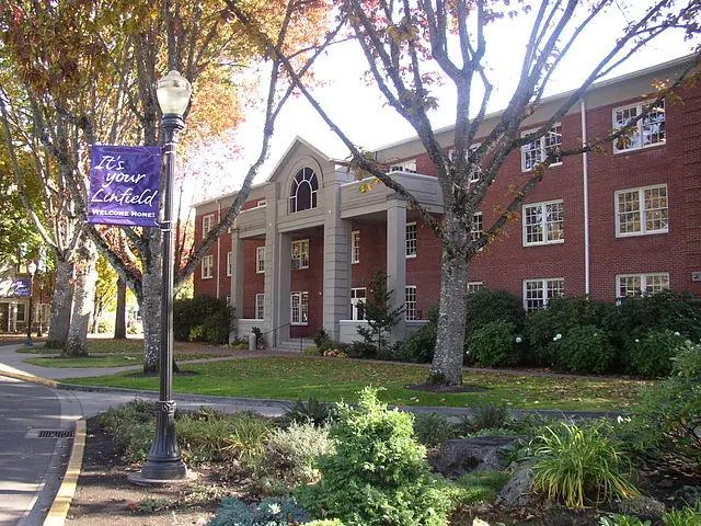 A red-brick building with white columns and arched windows is surrounded by trees with autumn leaves. A lamp post holds a purple banner reading, Its your Linfield, Welcome Home. A sidewalk curves beside the building.