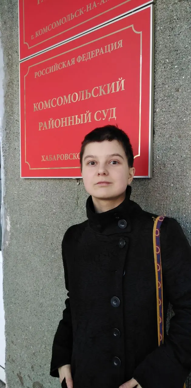 A person with short dark hair, wearing a black coat and carrying a colorful bag, stands in front of a wall with red signs in Russian indicating Komsomolsky District Court in Khabarovsk, Russia.