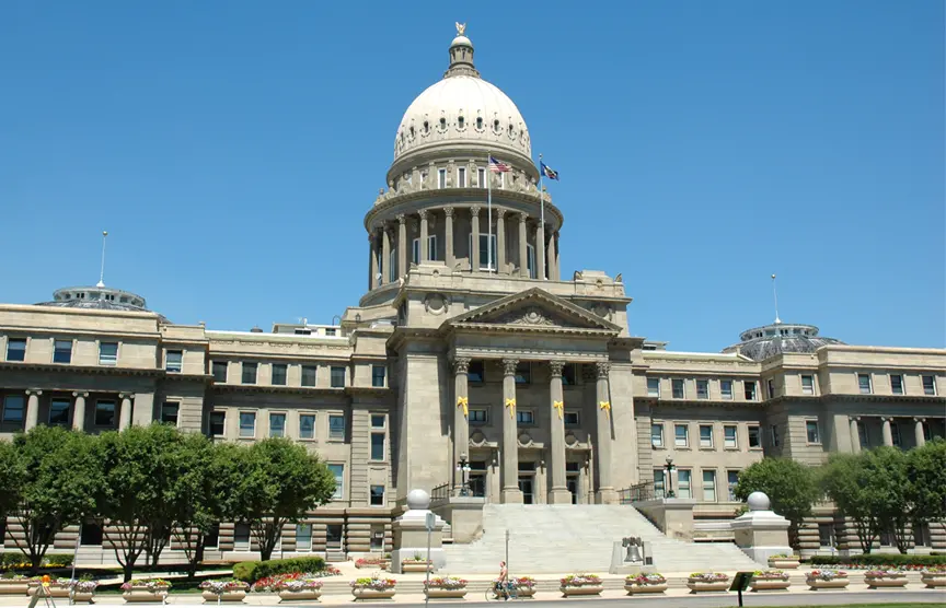The image shows a large, domed government building with columns at the entrance, wide steps, and a clear blue sky overhead. Manicured lawns and flower beds are visible in the foreground.