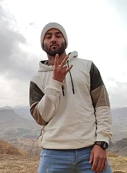 A man wearing a gray beanie, a light-colored hoodie with dark sleeves, and jeans stands outdoors, holding up three fingers with one hand. Mountains and a cloudy sky are visible in the background.