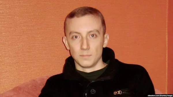 A man with short light brown hair, wearing a black jacket, sits in front of an orange textured wall and looks directly at the camera with a neutral expression.