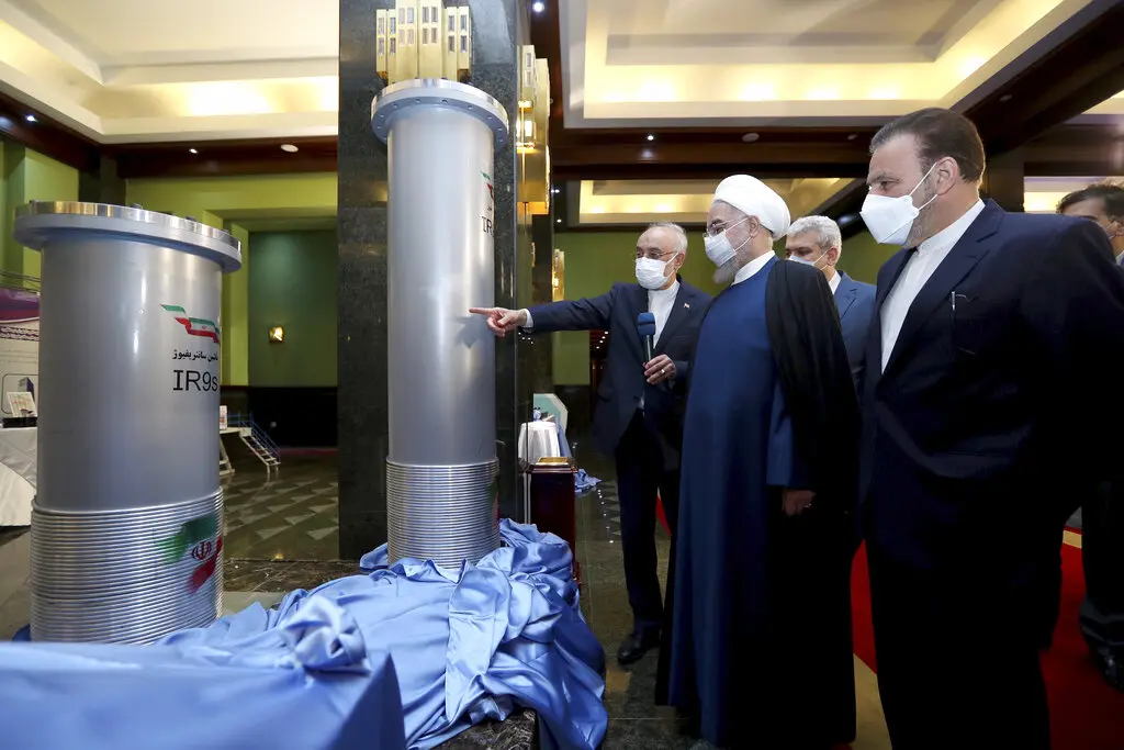 Several men wearing masks and formal attire examine and point at two large cylindrical machines on display, partially covered with blue cloth, inside a brightly lit room.