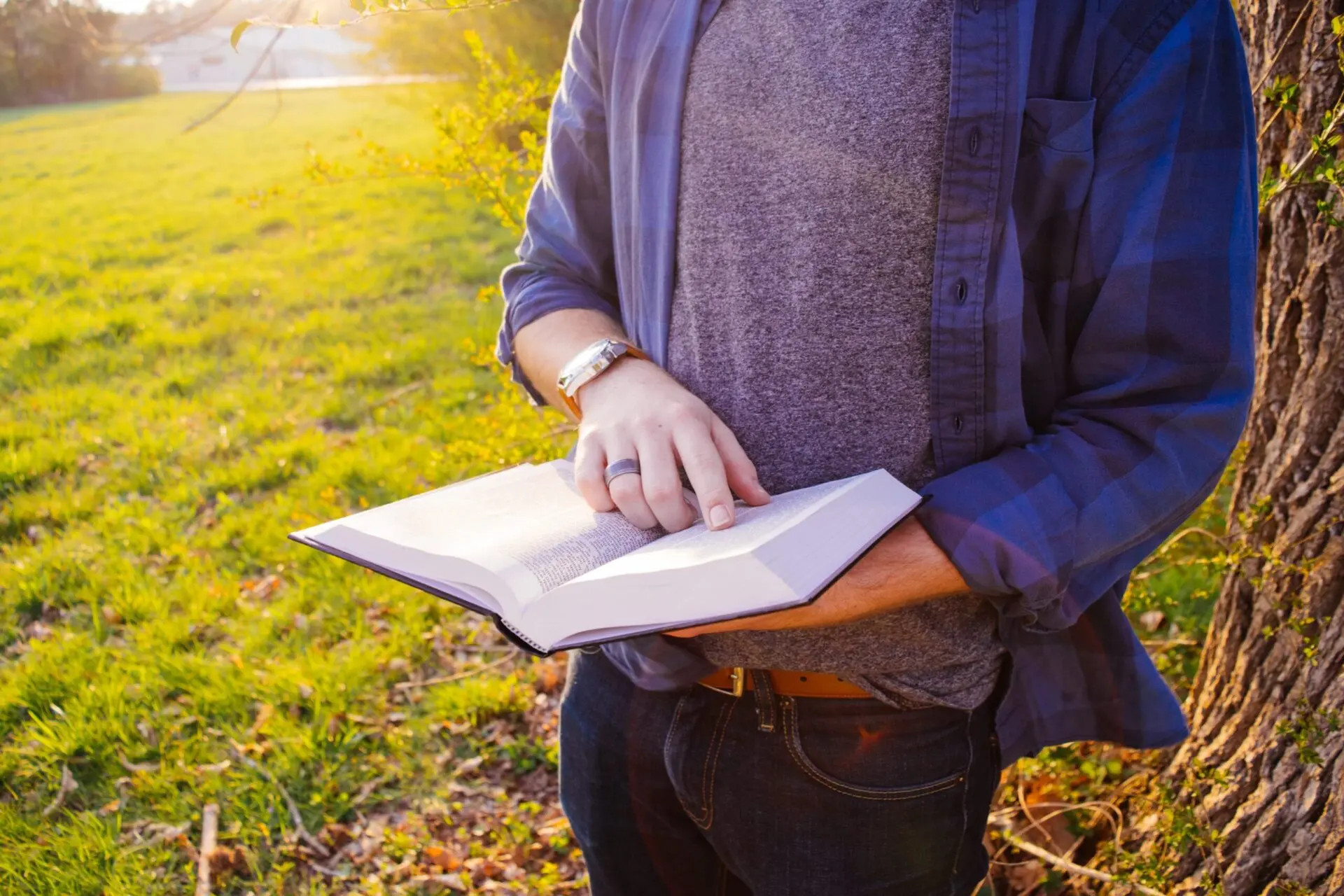 A person stands outside near a tree, wearing a blue shirt and jeans, holding an open book and reading with one finger pointing to the page. The background is grassy with sunlight shining.