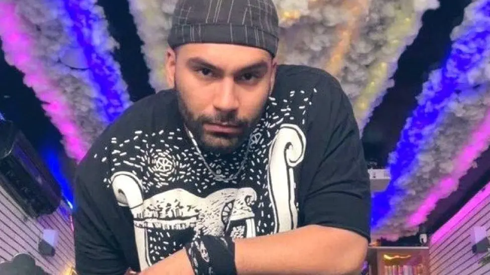 A man wearing a black and white graphic t-shirt and a striped beanie stares intently at the camera. The background features colorful lights and cotton-like decorations on the ceiling.