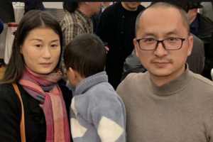 A woman with long dark hair and a scarf, a man with glasses and a gray sweater, and a child in a blue sweater stand together indoors, surrounded by other people in the background.