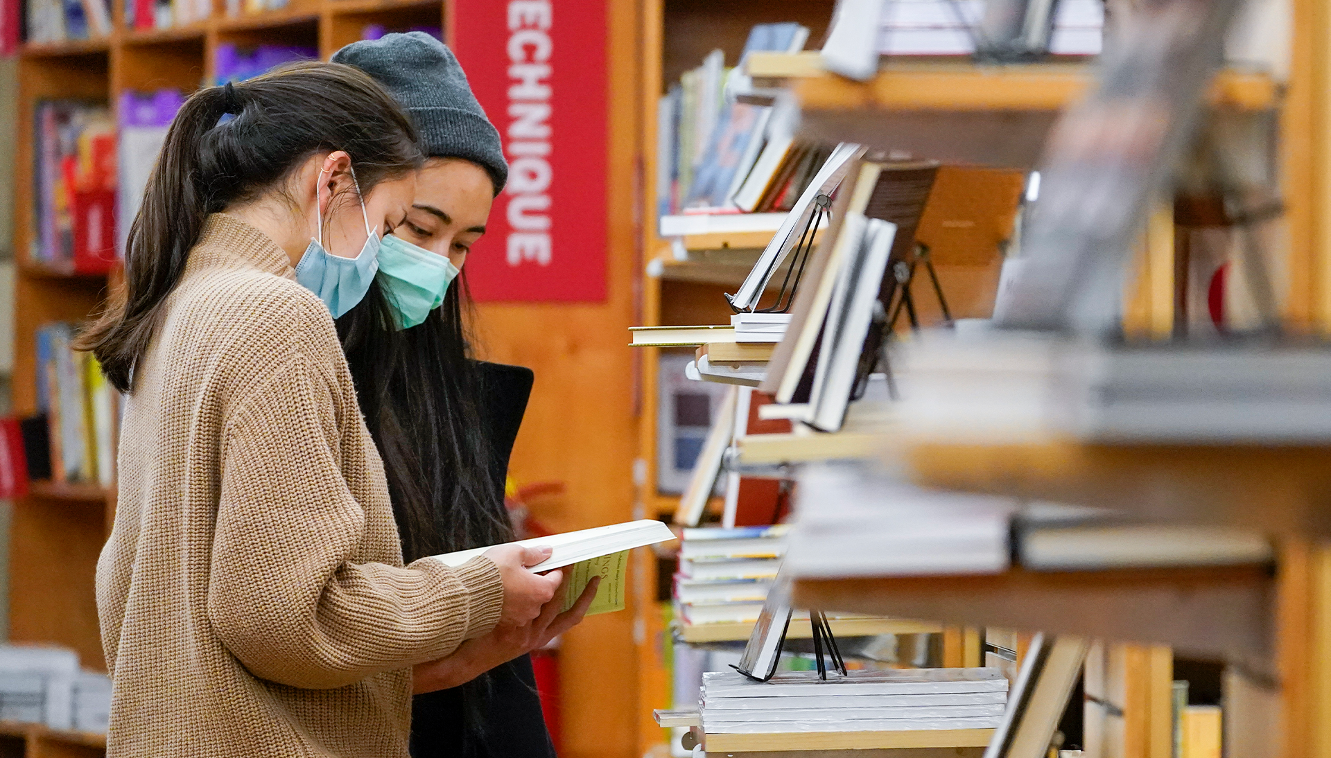 Two women wearing face masks look at a book together in a bookstore, standing near shelves filled with books. Members of the NYC Literary Action Coalition, they browse intently, with books and a red sign visible in the background.
