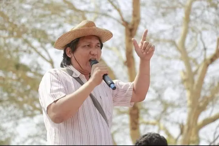 A person wearing a straw hat and striped shirt speaks into a microphone outdoors, raising one hand with a finger pointed upward. Blurry trees are visible in the background.