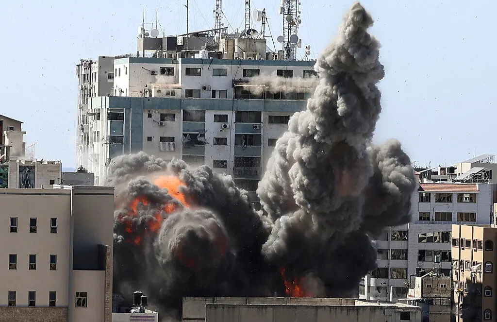 A large plume of dark smoke and flames rises from a building after an explosion in an urban area, with other buildings and communication towers visible in the background.