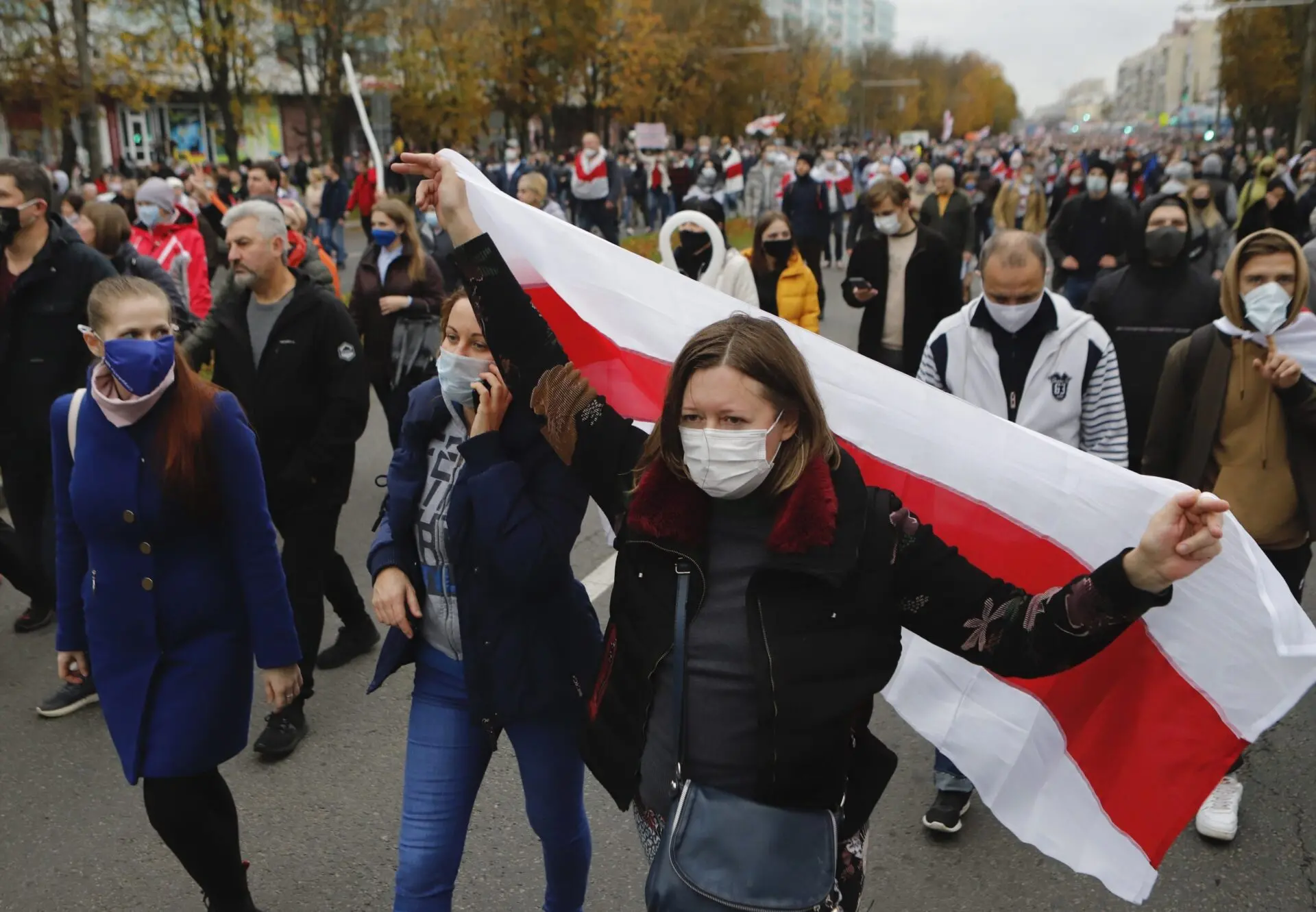 A group of people wearing masks march down a street; one woman in front holds up a red and white flag. Trees with autumn leaves and more protesters are visible in the background.