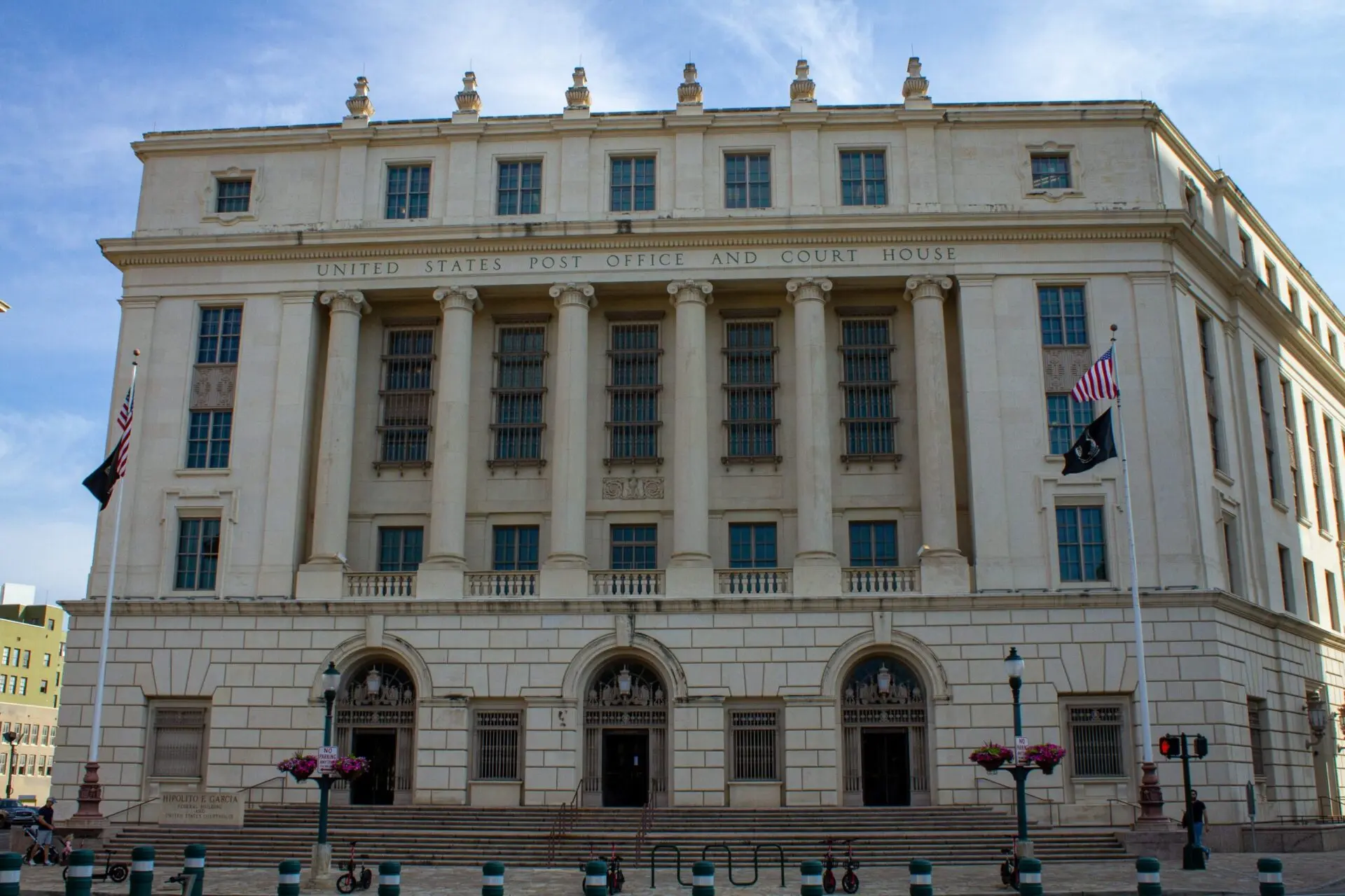 A large, historic building with tall columns and many windows, labeled United States Post Office and Court House on the front, with American flags and flower planters outside.