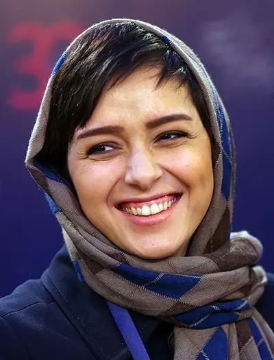A woman with short dark hair smiles warmly, wearing a brown and blue checkered headscarf against a blurred purple background.
