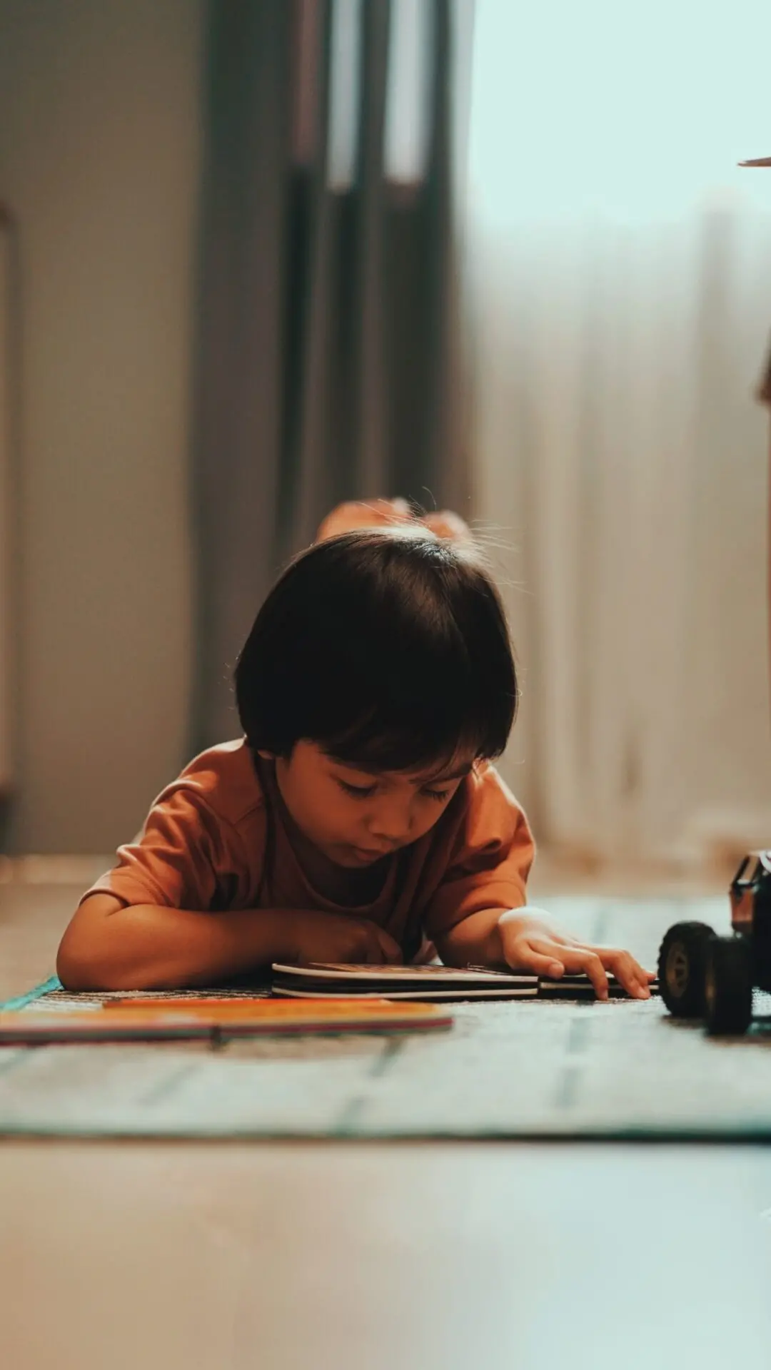 A young child with dark hair lies on the floor, intently drawing or coloring on paper. Nearby, a toy vehicle is visible. Soft natural light filters in from a window in the background.