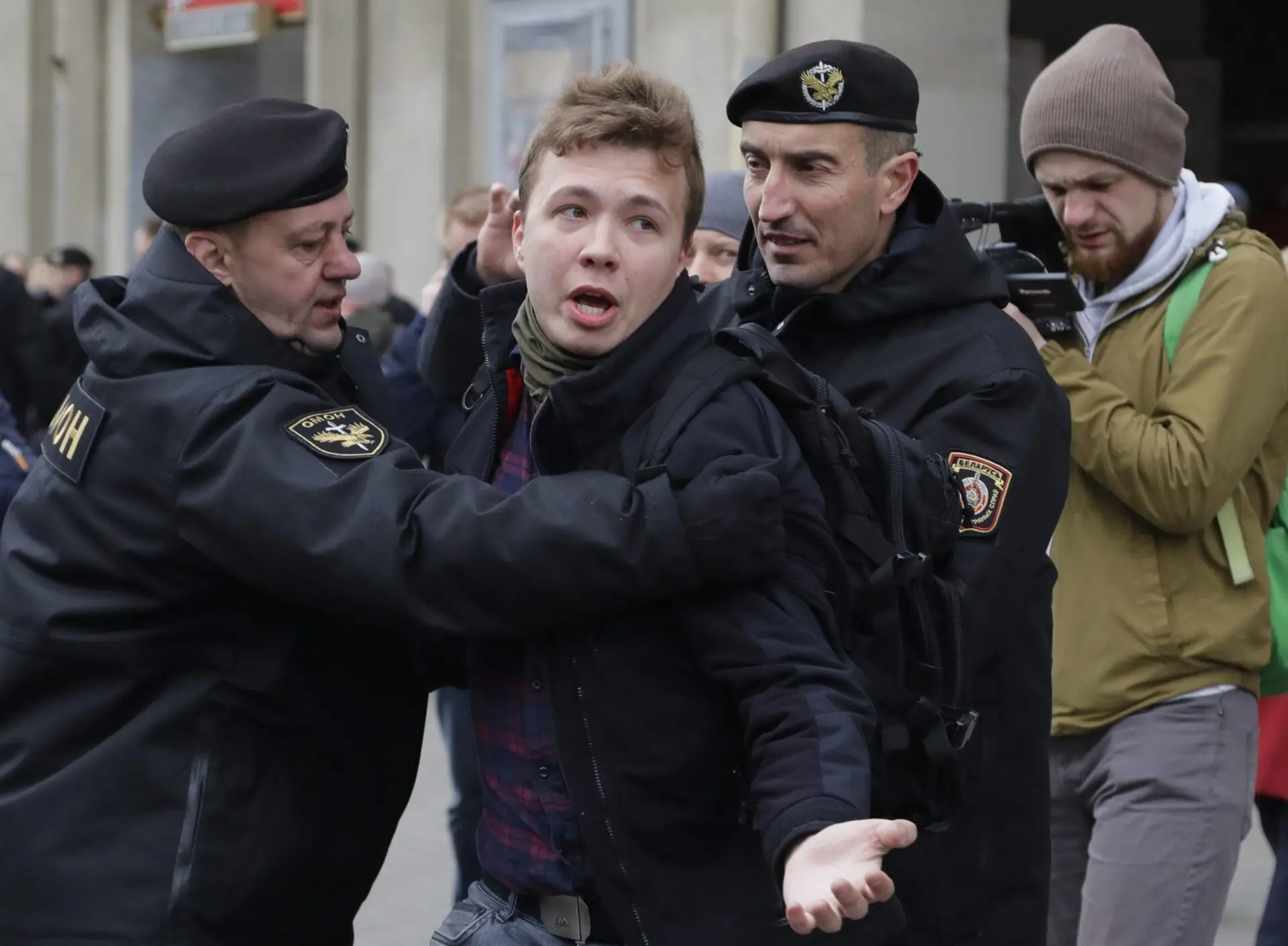 Two uniformed police officers detain a young man with a distressed expression. Another man with a camera stands in the background, capturing the scene outdoors.