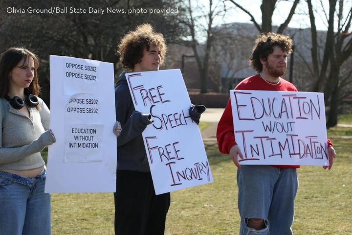 Three young adults stand outside holding protest signs that read “OPPOSE SB2012,” “Free Speech Free Inquiry,” and “Education w/out Intimidation.” Bare trees and a campus building are visible in the background.