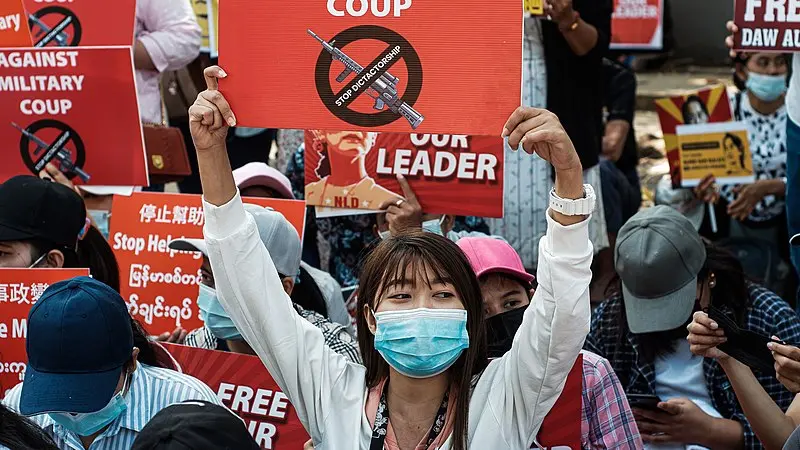A woman wearing a mask holds a red sign with a crossed-out gun symbol reading STOP MILITARY COUP at a protest, surrounded by others holding similar signs demanding freedom and opposing a coup.
