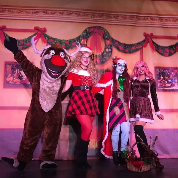 Four people in Christmas-themed costumes stand on stage: a reindeer mascot, a woman in a red plaid dress, a woman painted blue with a red cape, and a woman in a brown dress with pink hair. Festive decorations hang behind them.