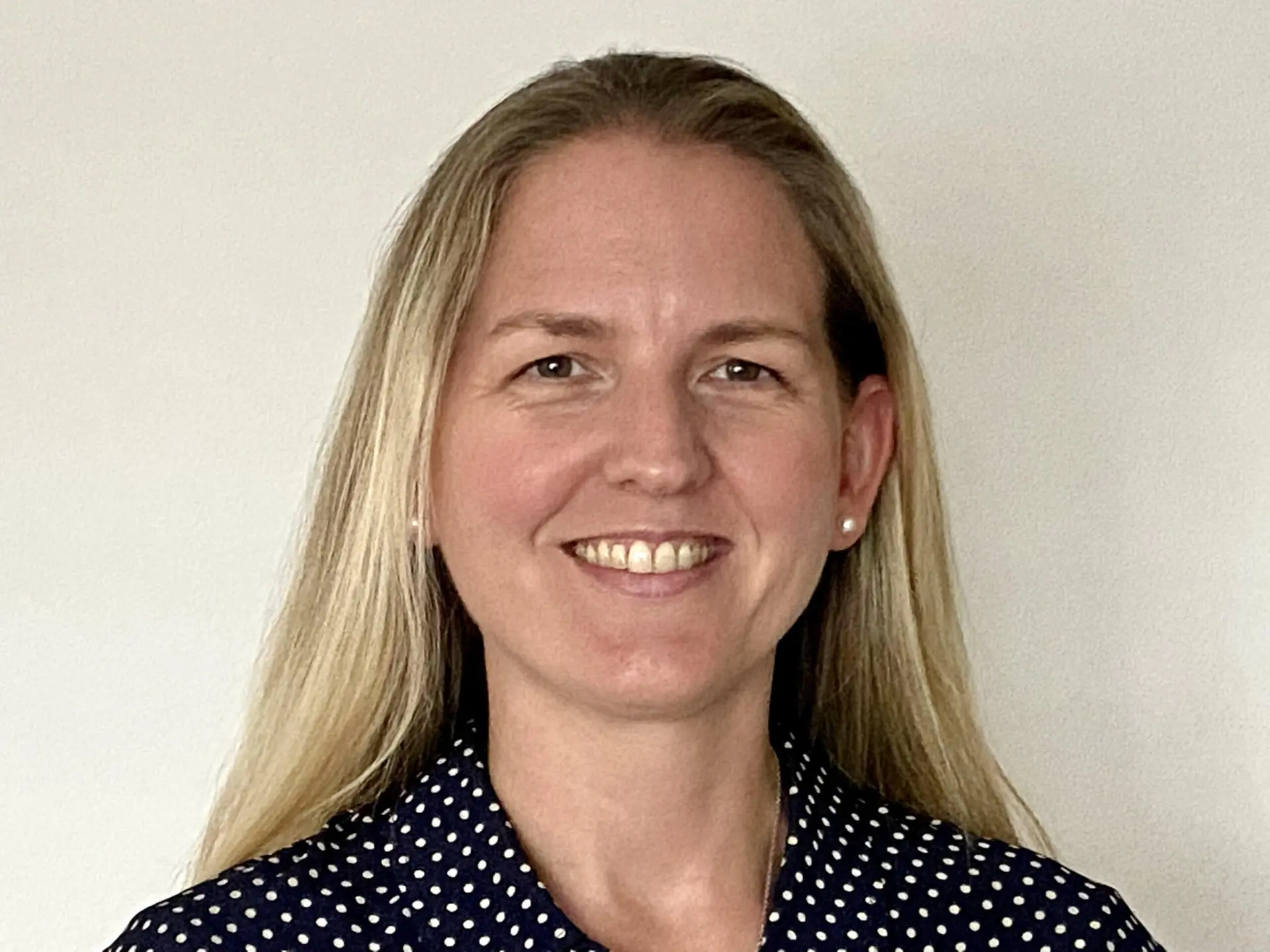 A woman with long blonde hair smiles at the camera. She is wearing a dark blue shirt with white polka dots and small stud earrings, standing against a plain light-colored background.